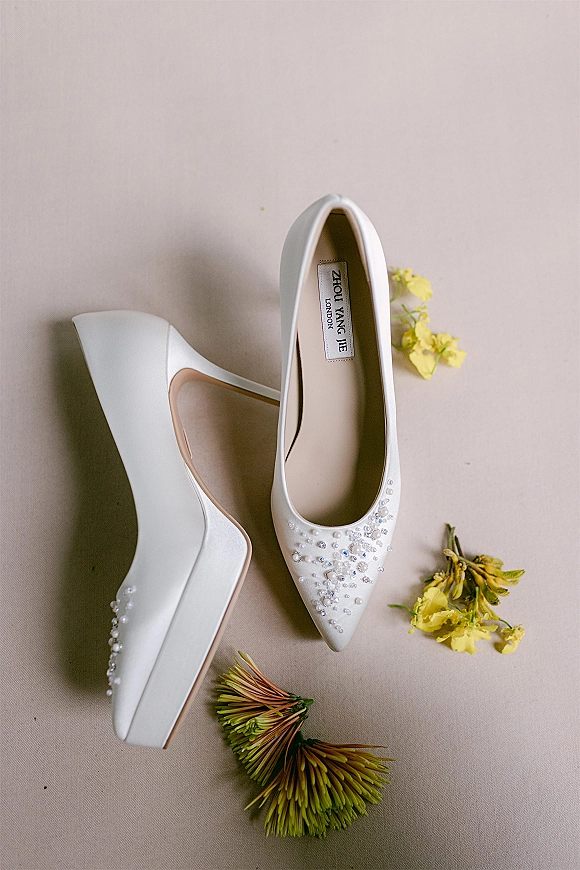 Bridal shoes in a flat lay, white wedding heels with pearl and crystal beading beside a yellow flower on neutral fabric backdrop