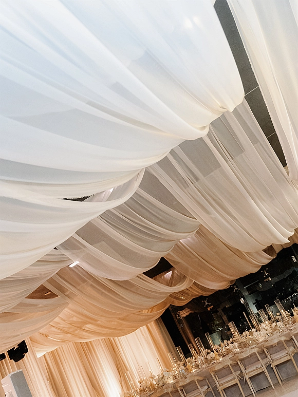 Ceiling drape decor with wedding ceiling draping, sheer white swags and uplighting over long banquet tables with florals and candles