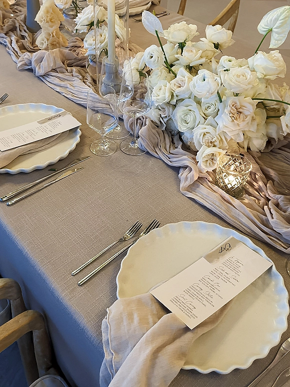 Reception tablescape with a wedding table centerpiece of white roses and ranunculus, gauze runner, taper candles, and scalloped plates in a neutral indoor space