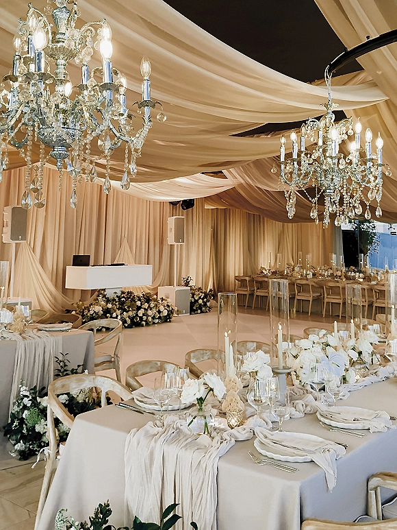 Reception tablescape with neutral wedding tablescape styling, white floral centerpiece and taper candles beneath draped fabric ceiling and chandeliers