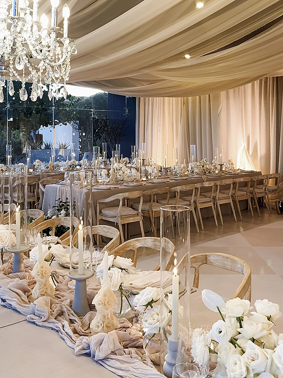 Reception tablescape with long banquet table wedding styling, taper candles and white florals under a crystal chandelier in a draped tent