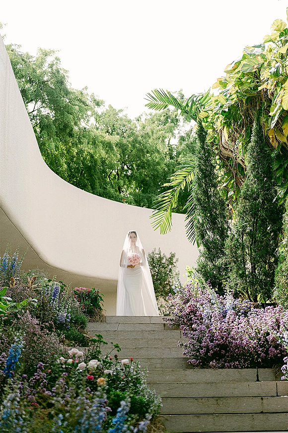 Bridal portrait of a bride holding bouquet of pink flowers, wearing a long veil and strapless white gown on garden stone steps by a curved wall