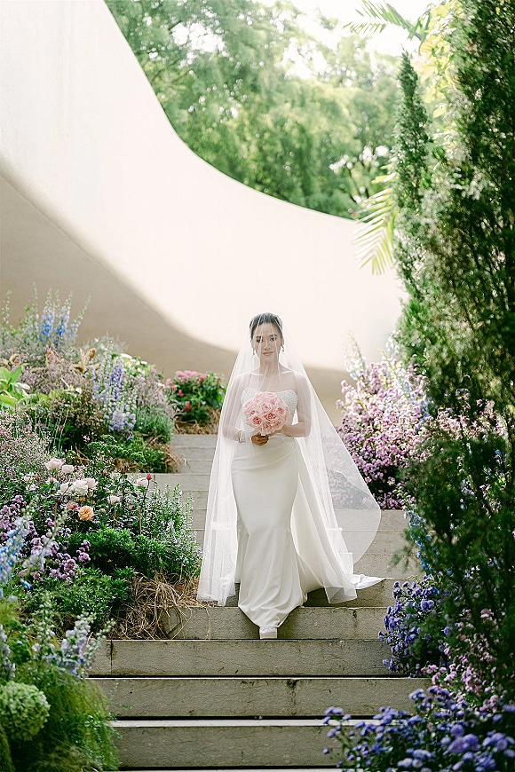 Bridal portrait of a bride holding bouquet of pink roses, wearing a veil and strapless dress on stone steps by garden flowers under a tent