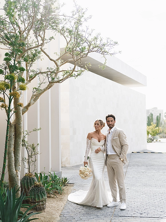 Couple portrait of bride and groom posing, bride holding bouquet, in front of a modern white building with desert cacti walkway