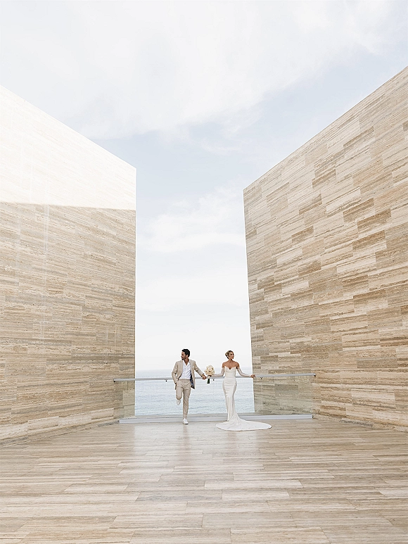 Couple portrait of bride and groom holding hands, bride with bouquet in strapless dress beside glass railing on ocean-view deck
