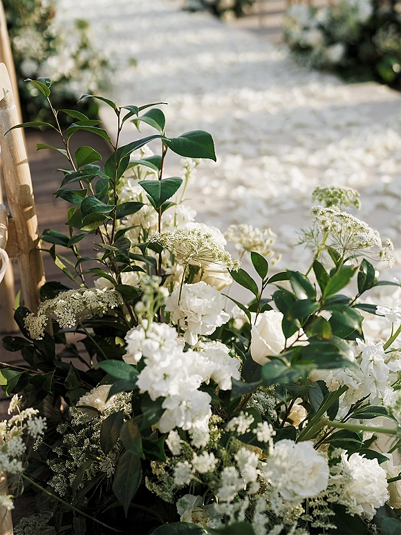 Ceremony aisle decor with white flower aisle decor and lush greenery lining an outdoor walkway, scattered petals leading past wooden chairs