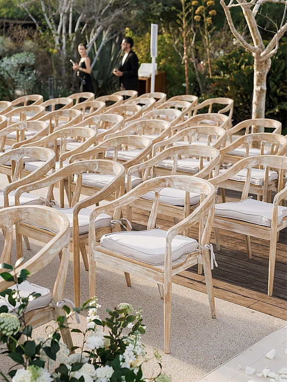 Ceremony seating with outdoor wedding seating, wood chairs and white cushions lining a petal-strewn aisle with floral clusters on a garden deck