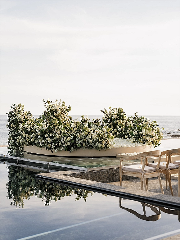 Ceremony altar decor with a floral ceremony arch of white florals and greenery, petal aisle and wooden chairs by an ocean-view pool