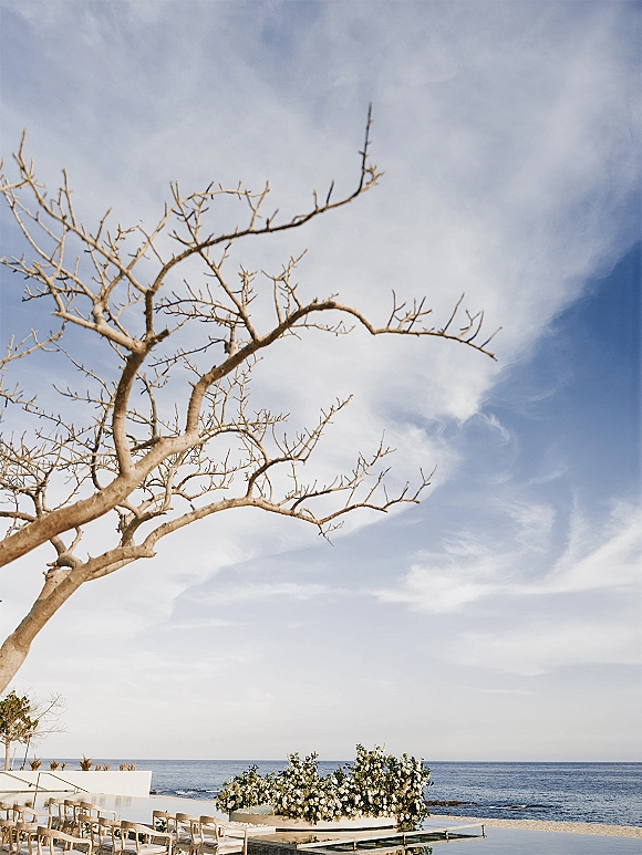 Ceremony setup for a beach wedding ceremony with white floral arrangements and greenery on a white aisle platform facing the ocean under blue sky