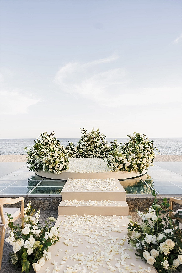 Ceremony setup with white floral arrangements and rose petals along a glass platform, facing the ocean horizon on the beach