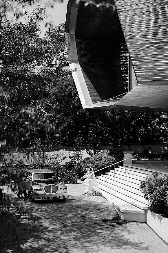 Wedding exit as the groom carries the bride in a veil toward a classic wedding car outside a modern building with stairs and trees
