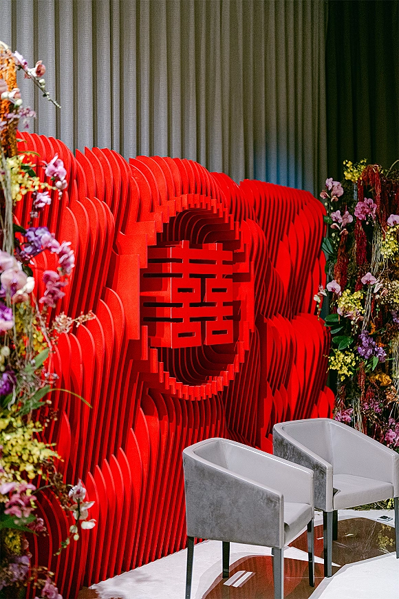Wedding ceremony backdrop with double happiness wedding decor, red sculptural stage, floral installations, and white chairs before pleated drapery