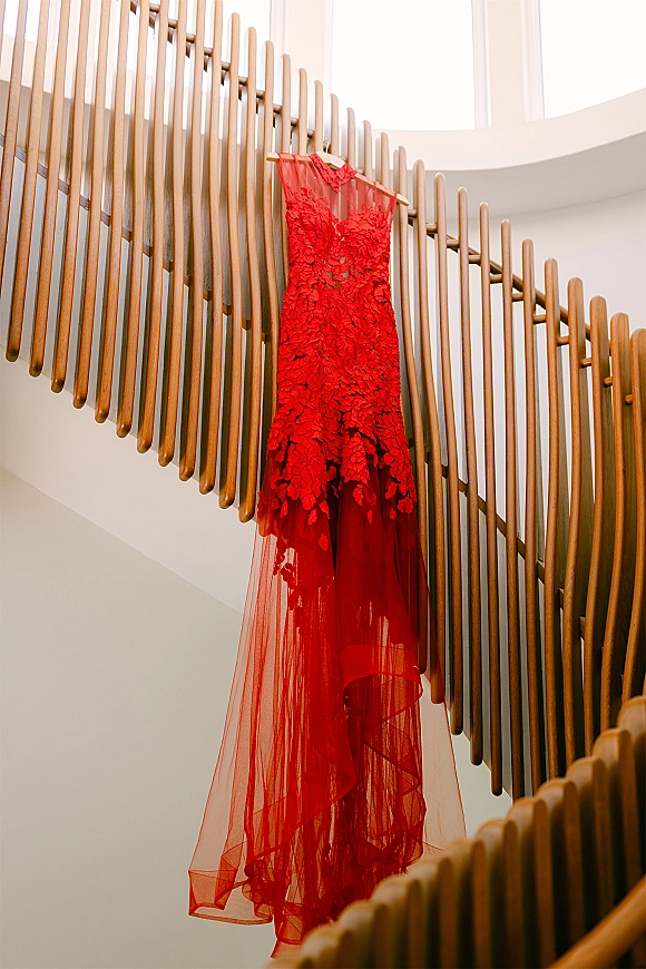 Red wedding dress with lace appliqué bodice and sheer tulle skirt on a wooden hanger by a curved staircase in natural light