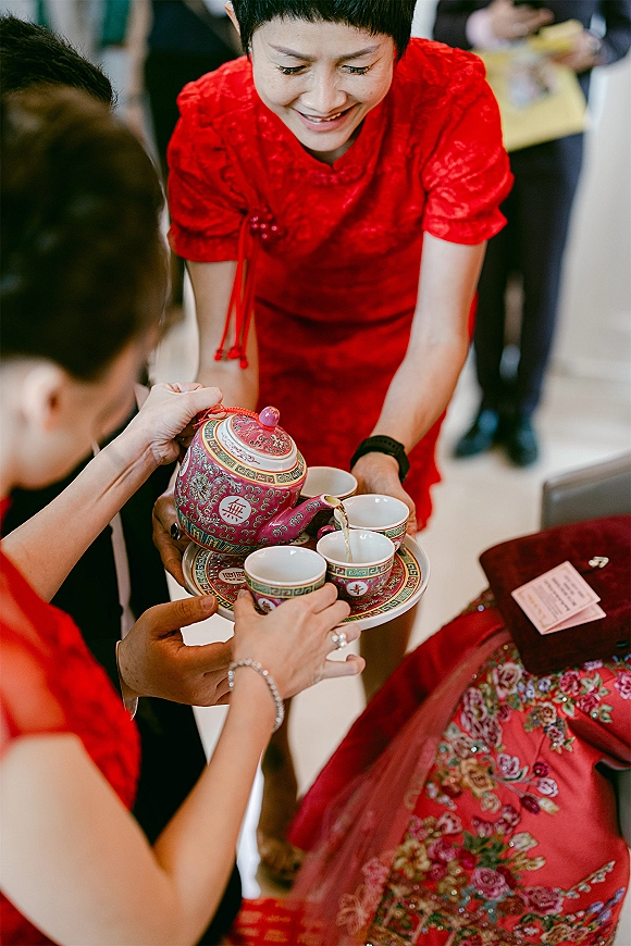 Tea ceremony with hands pouring from a porcelain teapot into teacups on a tray, red qipao accent amid indoor wedding guests