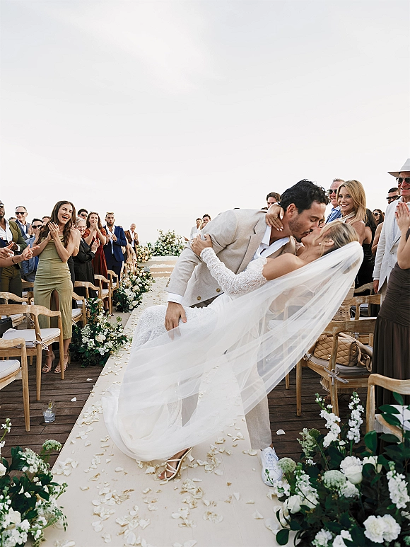 Wedding kiss portrait of a couple dip kiss in the aisle, bride’s veil flowing over lace dress as guests cheer on an outdoor deck