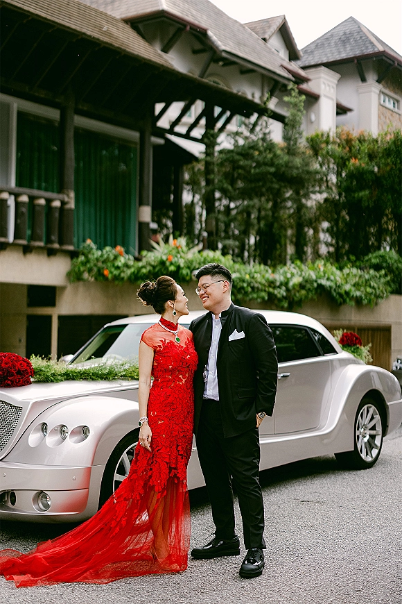 Couple portrait by a vintage luxury car, bride in red lace cheongsam and groom in black suit beside rose-decorated hood on street pavement