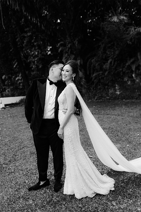 Couple portrait with groom kissing bride’s cheek as she smiles, in a strapless beaded wedding dress and long veil on garden lawn