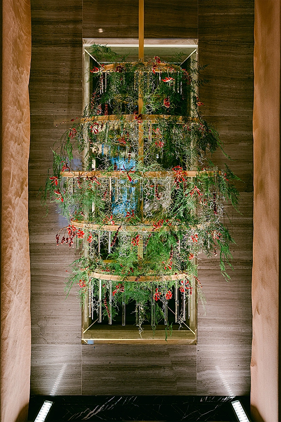 Hanging floral installation with wedding hanging greenery, red flowers, and glass tubes on a metal frame above a stone hallway