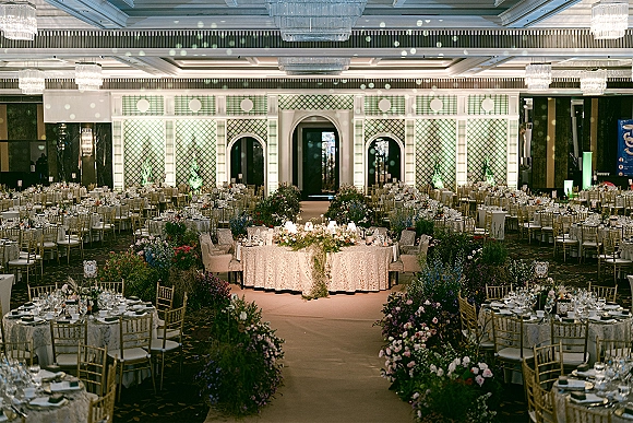 Wedding reception decor with lace-draped round tables, floral centerpieces and candlelight, gold chiavari chairs beneath ballroom chandeliers