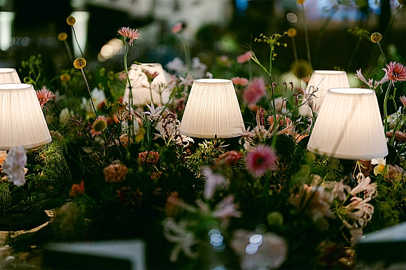 Reception tablescape with wedding table lamps glowing beside wildflower arrangements and greenery garland against dark garden foliage at night