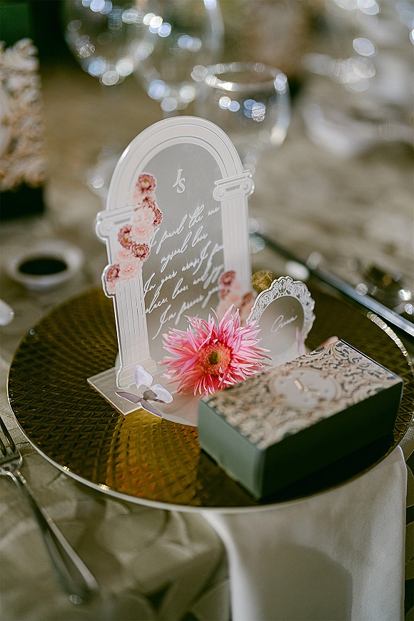 Wedding place card on an acrylic place card with pink floral illustration, set on a gold charger plate beside ring box and glassware