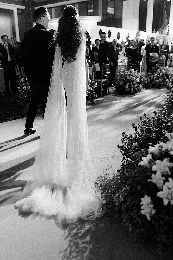 Wedding recessional as bride and groom walk down the aisle in a ceremony exit moment, her cape veil trailing past florals, guests in a columned indoor venue