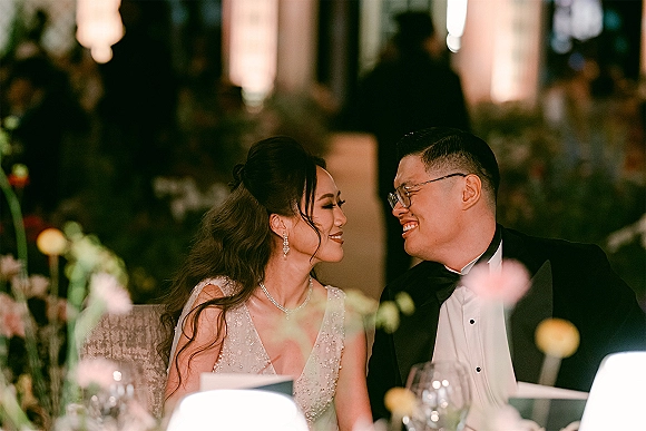 Couple portrait of bride and groom at table, smiling at each other in warm reception lighting with wine glasses and florals behind them