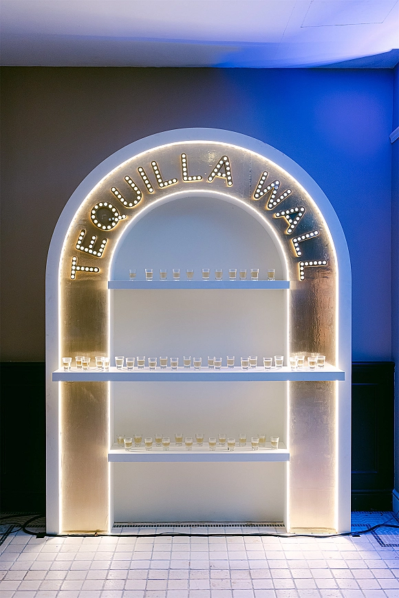 Tequila shot wall with an arched LED-lit display, marquee letters, and shelves of pre-poured shots and glasses against a painted wall