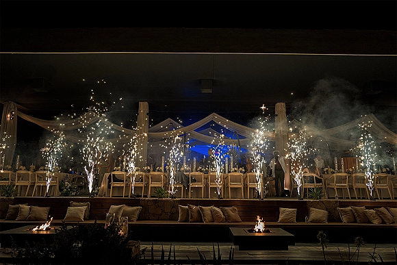 Wedding reception entrance with cold spark fountains under a draped fabric canopy, candlelit long tables and string lights on a smoky terrace at night