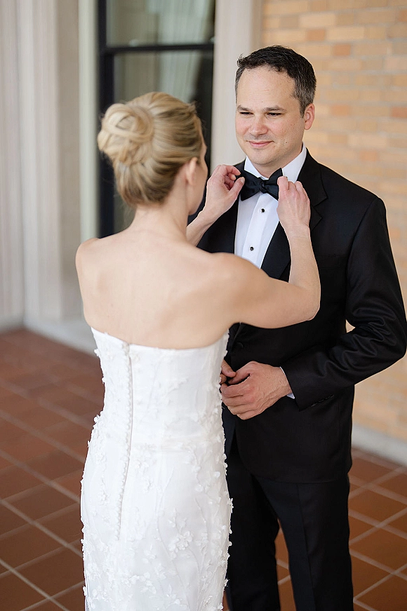 First look moment as the bride fixing bow tie on the groom in tuxedo, her strapless dress and updo beside a brick wall walkway