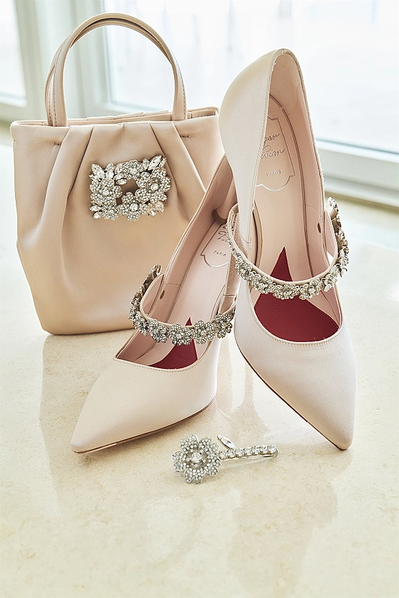 Bridal shoes with blush wedding heels and rhinestone straps arranged beside a bridal handbag on marble, lit by soft window light