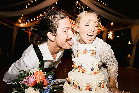 Wedding cake cutting as the bride and groom share a funny cake cutting photo beside a tiered floral buttercream cake under bistro lights in a tent