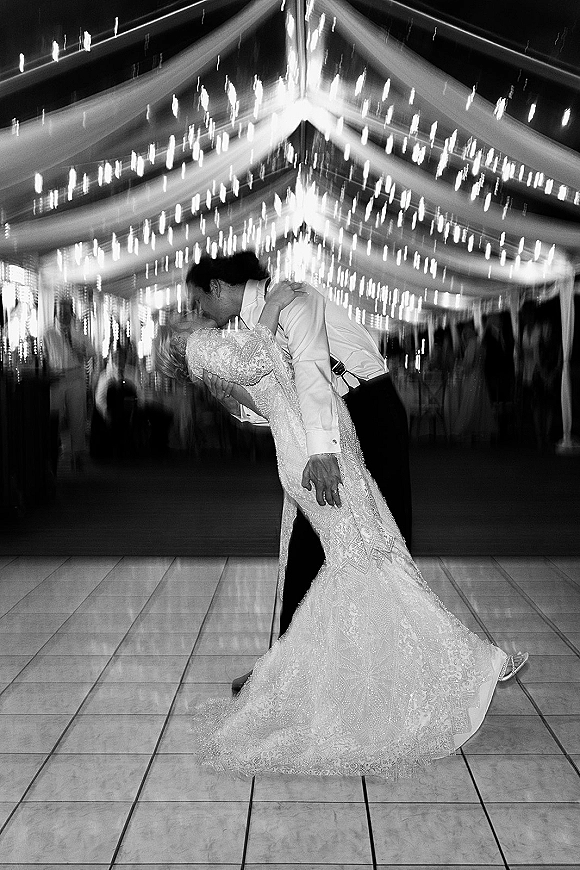 First dance kiss as groom dips bride in a lace long-sleeve wedding dress under string lights in a draped tent with reception crowd at night