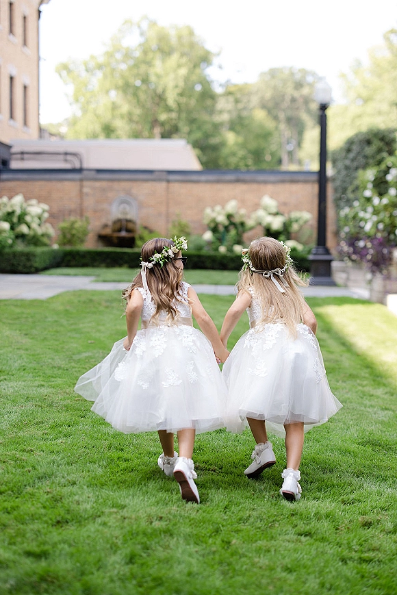 Flower girl moment as two flower girls holding hands walk away in matching tulle dresses across a grass lawn with garden hedges