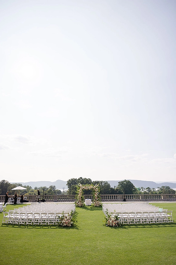 Ceremony setup with outdoor wedding ceremony seating, white folding chair rows and aisle florals leading to a floral arch on a terrace lawn overlook