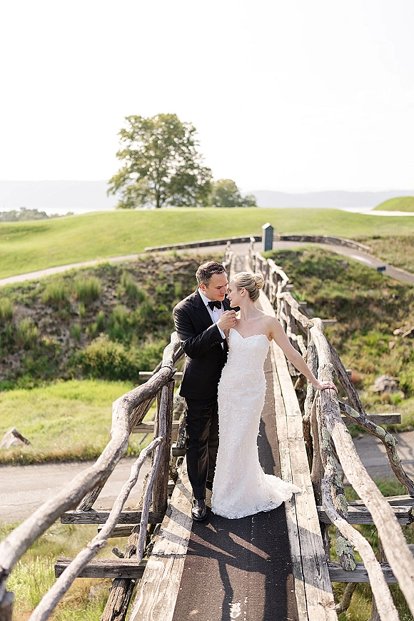 Couple portrait of bride in strapless lace wedding dress and veil as groom in black tuxedo kisses her forehead on a rustic wooden bridge