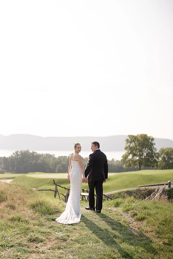 Couple portrait of bride and groom holding hands from behind on a grassy hill by a wooden fence, lake view, bride glancing over shoulder in veil