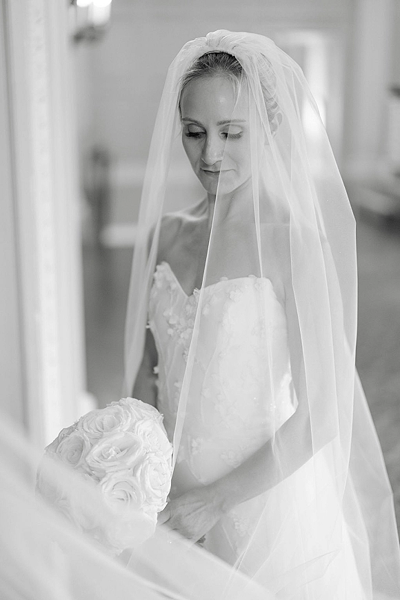 Bridal portrait in black and white of a bride with veil over her face, holding a rose bouquet in an indoor hallway with sconces