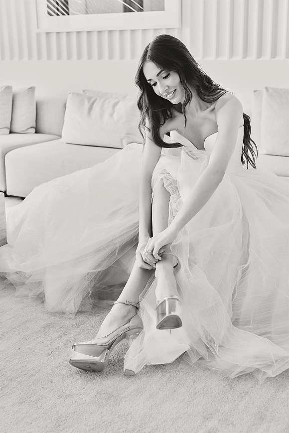 Bride getting ready, putting on wedding shoes while seated on a sofa in an indoor room, wearing a strapless dress with a tulle skirt