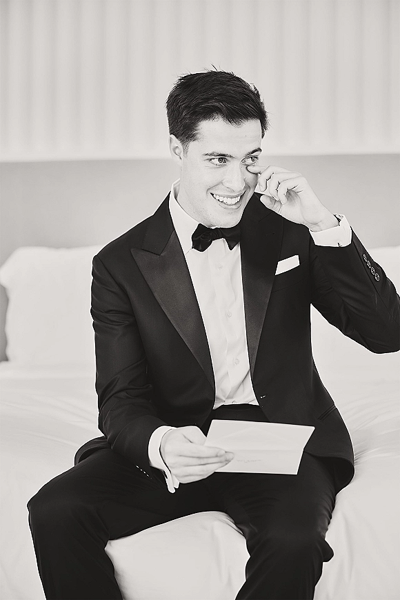 Groom portrait reading a letter in a black tuxedo, seated on a bed with white bedding and curtains, visibly moved with tears