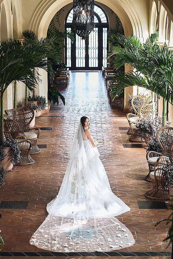 Bridal portrait of a bride in a cathedral veil looking back, her wedding dress train flowing down an arched terracotta hallway with petals
