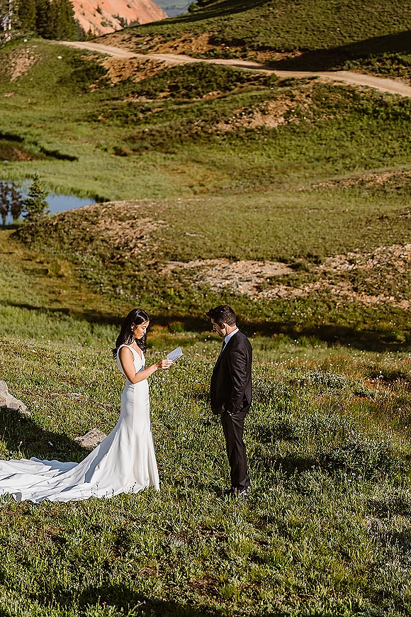 Wedding vows as bride reads from vow cards, long dress train flowing while groom in suit listens in a mountain meadow with wildflowers