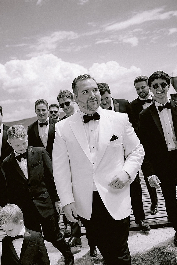 Groom portrait in a white tuxedo jacket with black bow tie, boutonniere, and sunglasses on a wooden walkway under cloudy sky