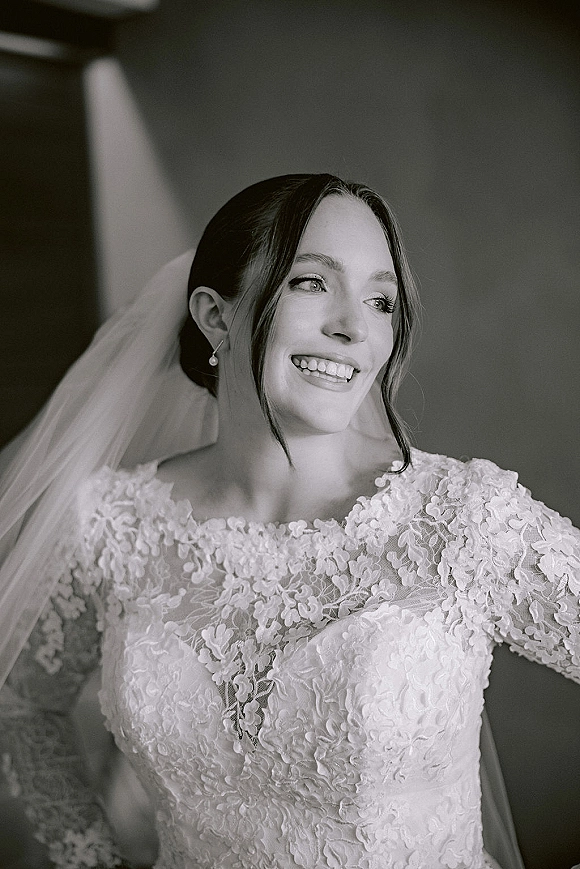 Bridal portrait in black and white of a smiling bride looking away in window light, wearing a lace long-sleeve dress and veil