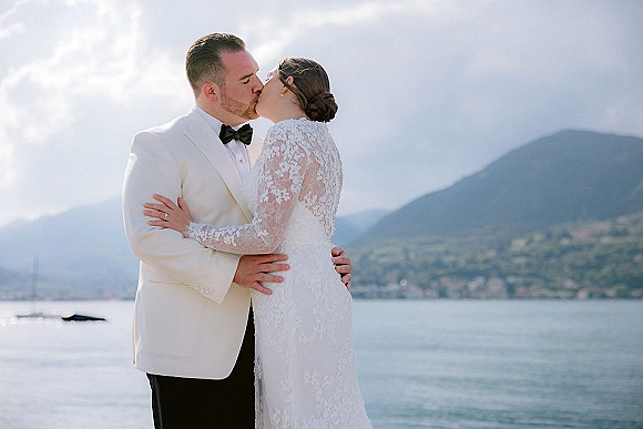 Wedding kiss between bride and groom kissing, her long sleeve lace dress visible, by a lakeshore with mountains under cloudy sky