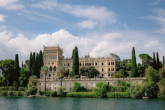Wedding venue exterior of a historic villa with arched windows and tower, set on a lakefront terrace with cypress trees under blue sky