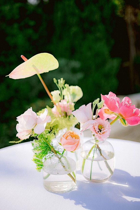 Wedding centerpieces featuring bud vase centerpiece designs with pastel roses, tulips and anthurium in clear glass vases on white linens outdoors