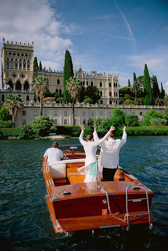Wedding getaway boat with newlyweds on boat waving from a wooden speedboat, Italian flag at stern, passing a lakefront villa with palms and cypress trees