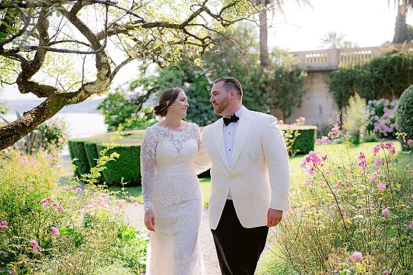 Couple portrait of bride and groom holding hands on a garden path, her long sleeve lace dress and his white dinner jacket by a lake