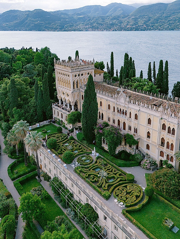 Wedding venue exterior with a historic villa façade, ornate arches, and terrace overlooking a parterre garden, lake, and mountains beyond